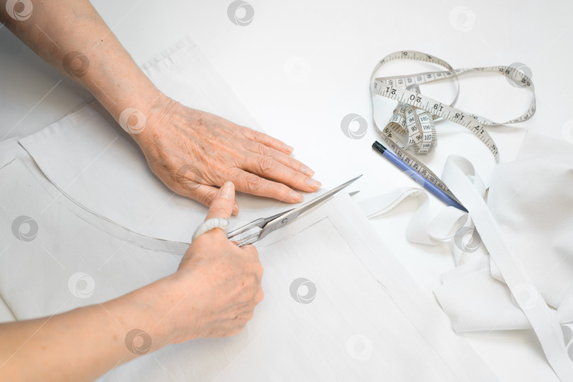 Скачать Руки пожилой женщины режут ножницами белую ткань, вид сверху. Карандаш и рулетка на белом столе. Старшая женщина-портниха. Работа на пенсии или хобби. фотосток Ozero