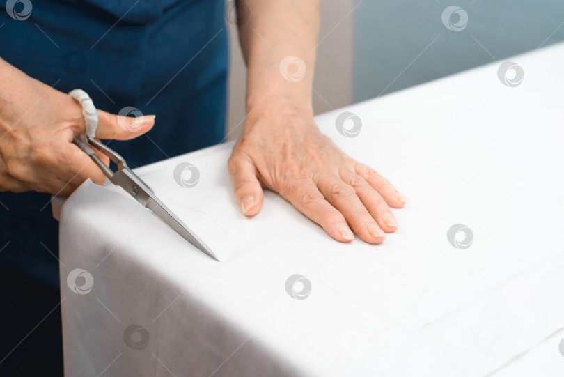 Скачать Пожилая женщина-портниха режет ножницами белую ткань крупным планом. Избирательный акцент на ножницах фотосток Ozero