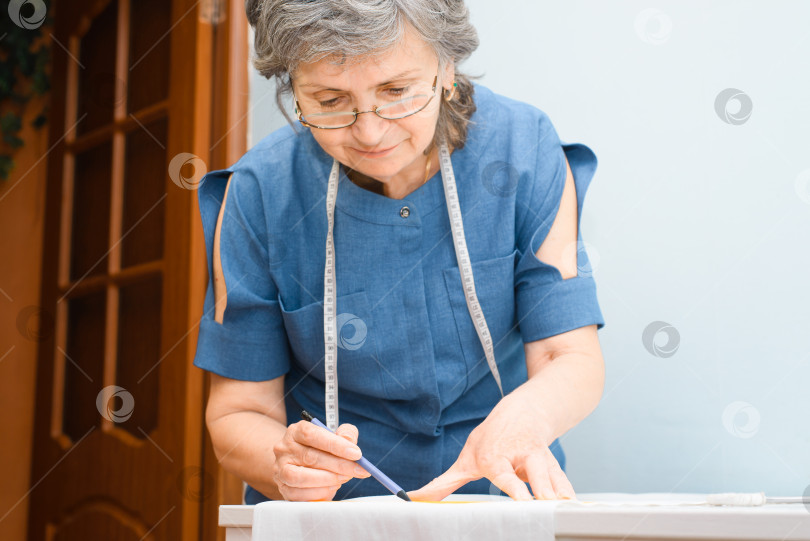 Скачать Пожилая женщина-швея, разрабатывающая одежду, обрезанное изображение. Улыбающаяся пожилая женщина в очках делает карандашный набросок на ткани. Хобби на пенсии, работа на дому. Выборочное сосредоточение внимания на руках. фотосток Ozero