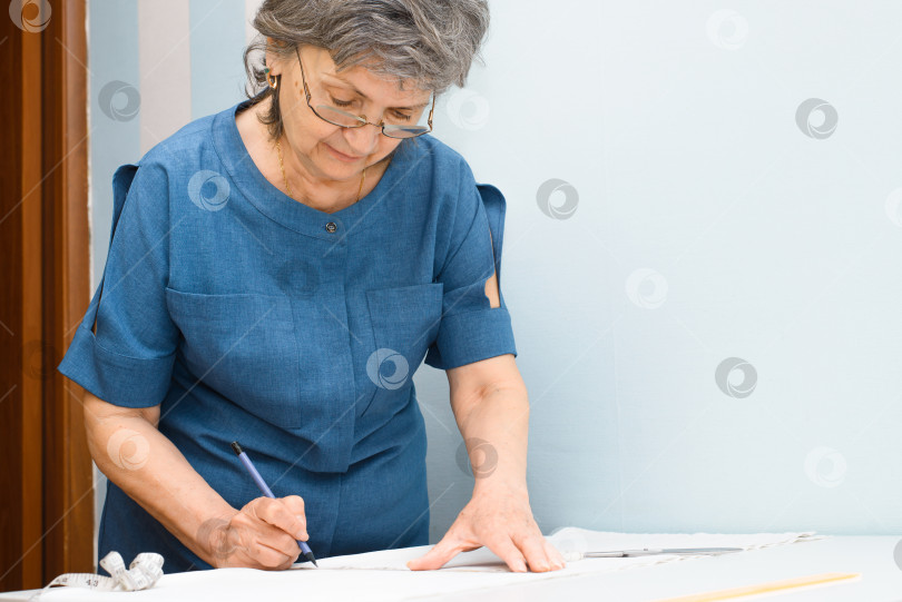 Скачать Пожилая женщина-швея, разрабатывающая дизайн одежды в помещении. Улыбающаяся пожилая женщина в очках делает набросок одежды карандашом на ткани. Избирательный фокус. фотосток Ozero