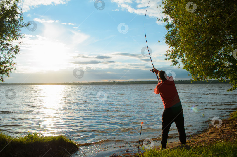 Скачать Рыбак на озере. Мужчина забрасывает спиннинг, вид сзади. Рыбалка на закате. Заходящее солнце, берег озера в обрамлении деревьев. фотосток Ozero