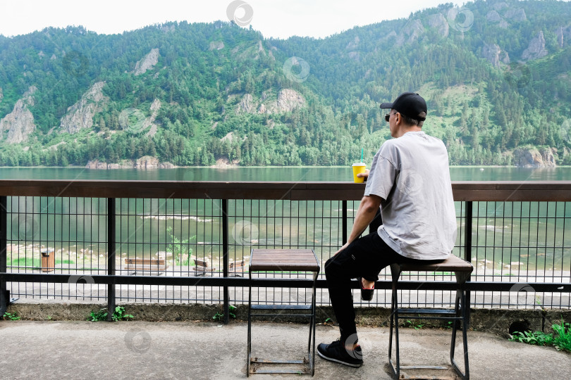 Скачать Вид сзади на мужчину-путешественника, сидящего на деревянной скамейке и наслаждающегося удивительной природой в горах. Мужчина пьет кофе на открытом воздухе по утрам. фотосток Ozero