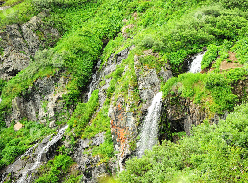 Скачать Каскад из нескольких небольших водопадов стекает со скалы, поросшей травой и мелким кустарником. фотосток Ozero