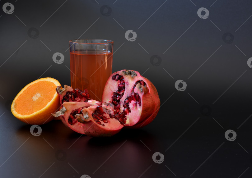 Скачать Высокий стакан с апельсиновым соком и гранатом на черном фоне, рядом с половинками спелых фруктов. фотосток Ozero