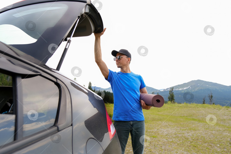 Скачать Мужчины стоят возле машины с открытым багажником, планируя заняться йогой на коврике. Йога на открытом воздухе в горах фотосток Ozero