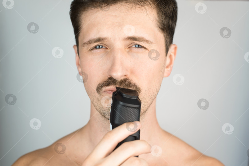 Скачать Портрет мужчины, подстригающего усы в помещении, крупным планом. Молодой усатый мужчина с современным электрическим триммером для бороды. фотосток Ozero