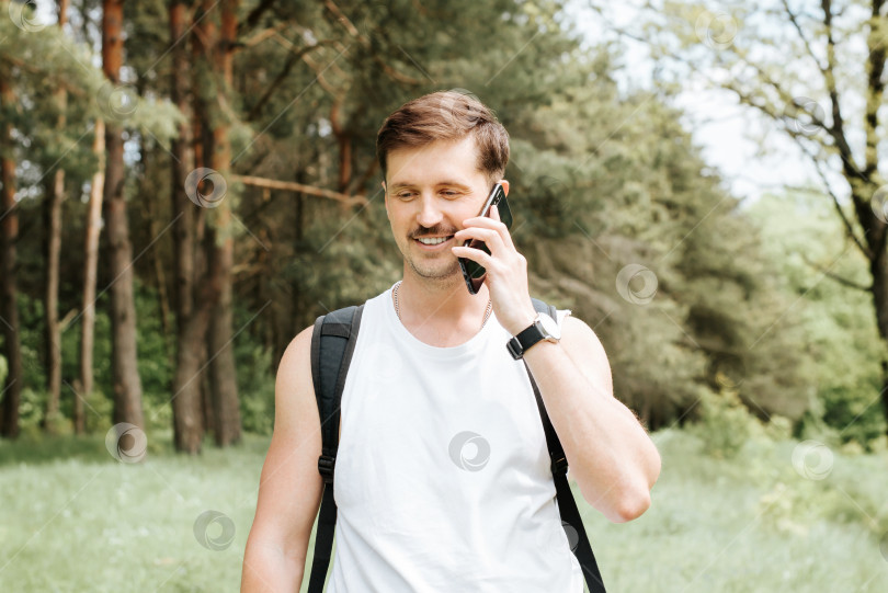 Скачать Портрет улыбающегося молодого человека, разговаривающего по мобильному телефону во время прогулки по лесу. Счастливый человек с рюкзаком отправляется в поход на природу, приятный разговор на свежем воздухе. фотосток Ozero