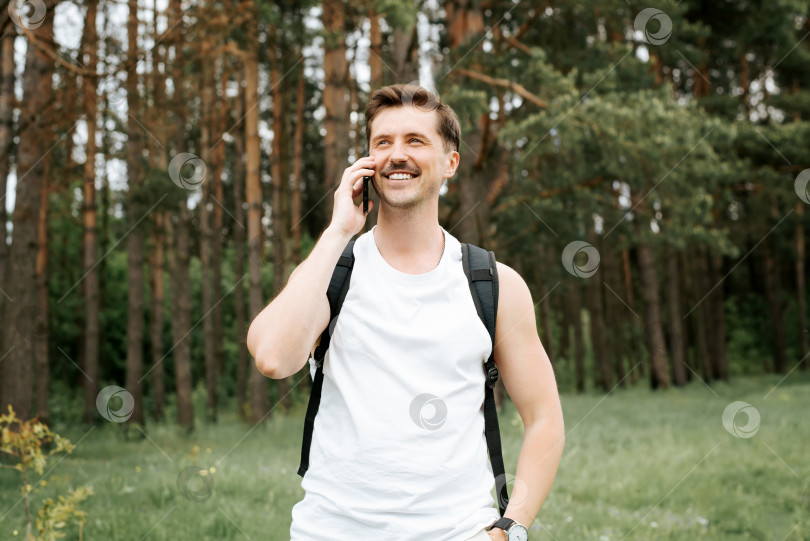 Скачать Счастливый мужчина разговаривает по мобильному телефону во время прогулки по лесу. Спортивный путешественник с рюкзаком, улыбающийся и держащий в руках телефон, на фоне соснового леса на открытом воздухе. Образ жизни красивого молодого человека фотосток Ozero