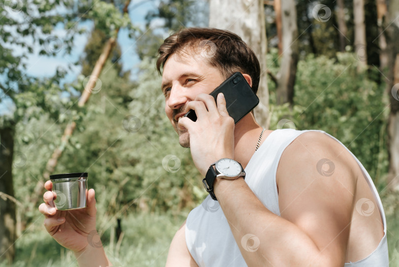 Скачать Человек отдыхает на природе. Улыбающийся молодой человек пьет чай в лесу и разговаривает по мобильному телефону. Счастливый путешественник отдыхает во время пешего похода на свежем воздухе. фотосток Ozero