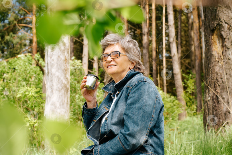 Скачать Портрет счастливой зрелой женщины, отдыхающей на свежем воздухе в лесу. Улыбающаяся пожилая женщина в очках держит кружку-термос с горячим чаем и смотрит на небо. Концепция отдыха, рекреации на природе. фотосток Ozero