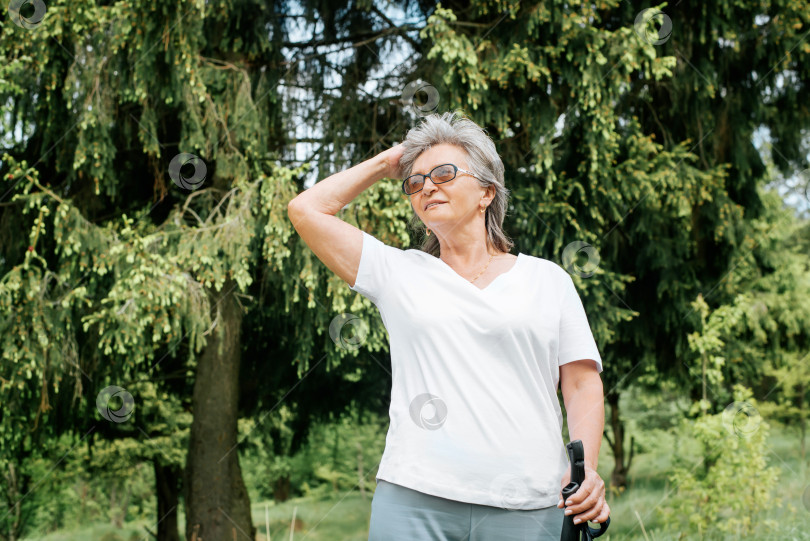 Скачать Пожилая женщина прогуливается на свежем воздухе. Улыбающаяся зрелая женщина, стоящая с палками для скандинавской ходьбы в лесу летним днем. Веселая пенсионерка наслаждается прогулкой, поправляя волосы. Активный образ жизни на пенсии фотосток Ozero
