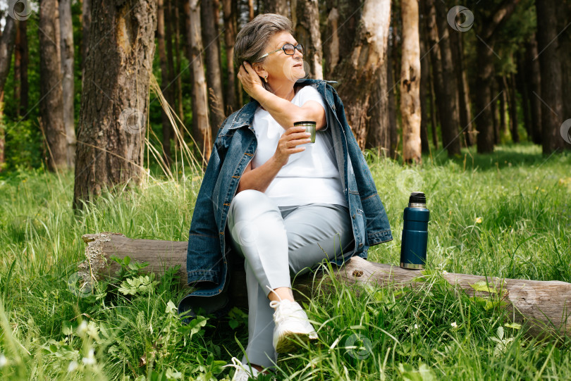Скачать Зрелая женщина отдыхает на природе. Пожилая женщина сидит на бревне в лесу и пьет чай из термоса на открытом воздухе. Пенсионер отдыхает в кемпинге, наслаждаясь отдыхом. фотосток Ozero