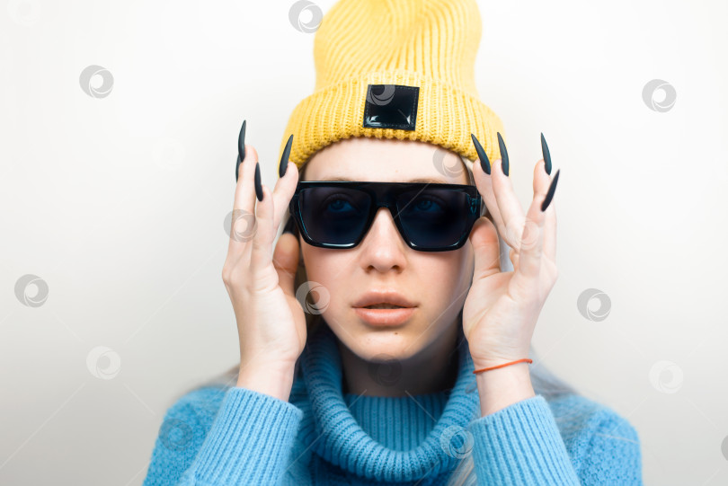 Скачать Молодая женщина в черных очках, желтой шапочке и вязаном свитере на светлом фоне. Подростковый наряд фотосток Ozero