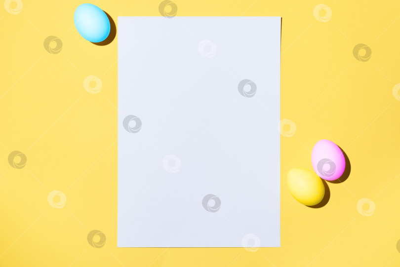 Скачать Пасхальный макет. Белый чистый лист бумаги и пасхальные разноцветные яйца на желтом фоне, ровно разложенные. Поздравительная открытка с местом для текста фотосток Ozero