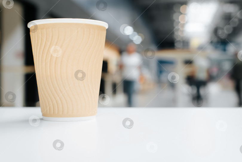 Скачать Одноразовый бумажный стаканчик с кофе стоит на белом столе в кафе, расфокусированный световой фон общественного места, в помещении. Выборочный акцент на чашке с напитками, копирование пространства. Вид сбоку на уровне объекта фотосток Ozero