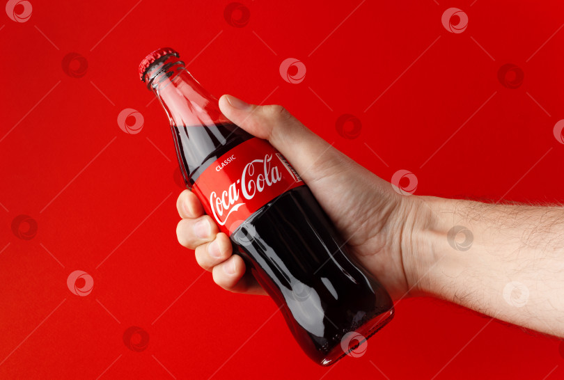 Скачать БАТУМИ, ГРУЗИЯ, 15.03.2023: Холодная стеклянная бутылка coca-cola в руке на красном фоне. фотосток Ozero