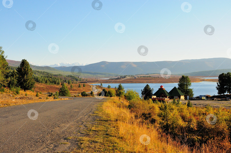 Скачать Полевая дорога спускается с холма к домам на берегу живописного озера с видом на заснеженные горы. фотосток Ozero