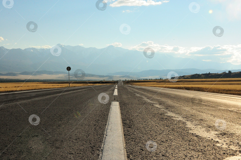 Скачать Белая разметка полосы движения на прямой асфальтированной дороге, идущей через степь к высоким горам. фотосток Ozero