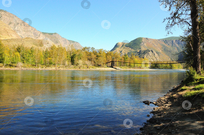 Скачать Спокойная гладь горной реки и подвесной мост через нее, окруженный высокими горами. фотосток Ozero