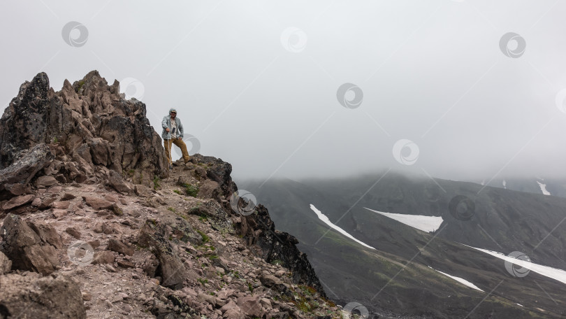 Скачать Мужчина с треккинговой палкой стоит на вершине скалы. Потрескавшиеся камни на крутых склонах, лишенных растительности фотосток Ozero