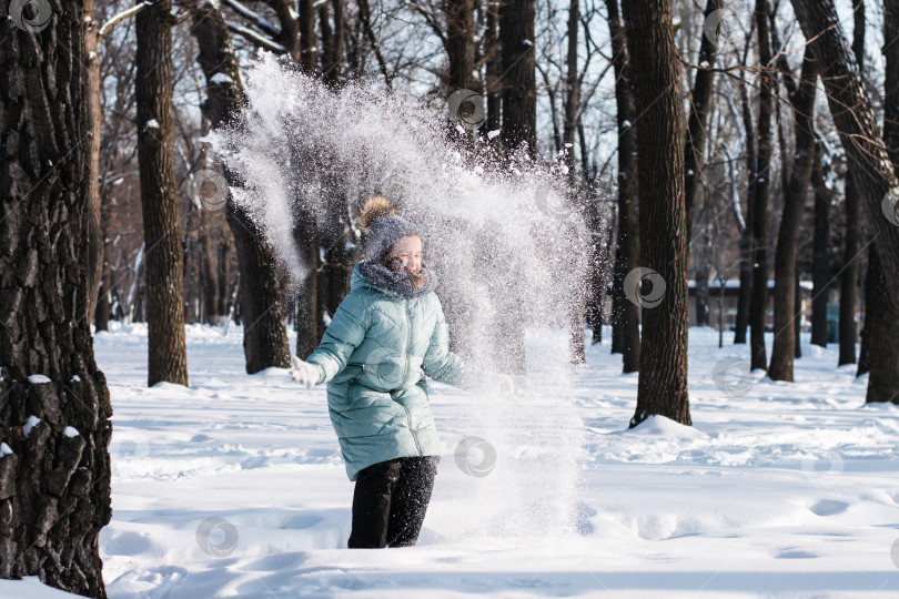 Скачать Счастливая девушка в теплой одежде разбрасывает снег в зимнем лесу. Вал фотосток Ozero