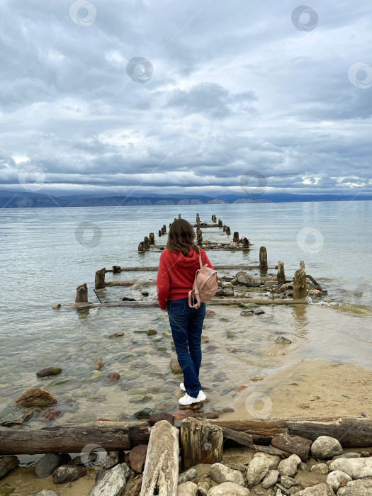 Скачать Молодая женщина с рюкзаком стоит возле озера Байкал, на старом разрушенном пирсе. фотосток Ozero