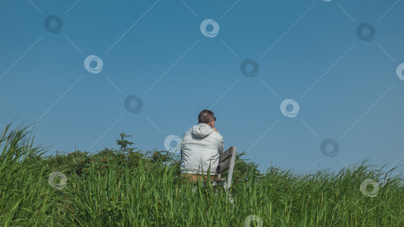 Скачать На зеленом лугу, среди сочной травы, стоит скамейка. Мужчина сидит на фоне ясного голубого неба фотосток Ozero
