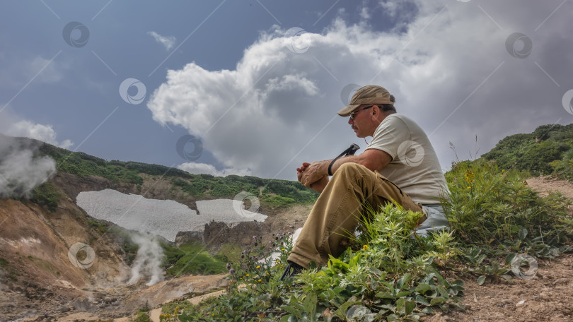 Скачать Мужчина с треккинговой палкой в руках сидит, отдыхая, на холме среди зеленой травы. фотосток Ozero