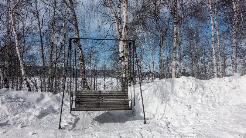 Скачать Деревянные некрашеные качели-скамейка стоят в заснеженном парке. фотосток Ozero
