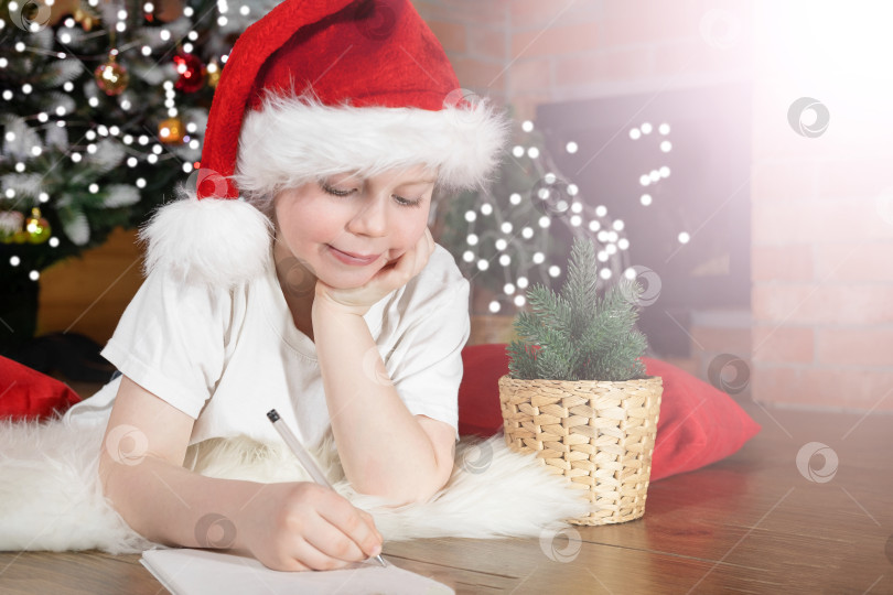 Скачать Счастливого Рождества, дети. Мальчик в шляпе Санта-Клауса пишет письмо Санте под рождественской елкой с рождественскими гирляндами, возле камина. Семейное Рождество. фотосток Ozero