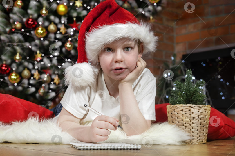 Скачать Счастливого Рождества, дети. Мальчик в шляпе Санта-Клауса пишет письмо Санте под рождественской елкой с рождественскими гирляндами, возле камина. Семейное Рождество. фотосток Ozero