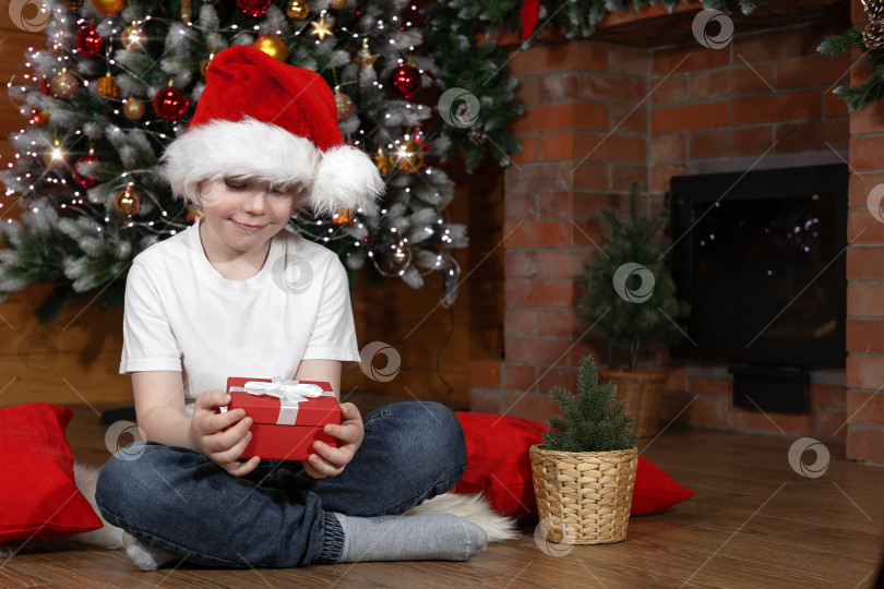 Скачать Рождественские дети. Мальчик в шапке Санта-Клауса с подарком от Санта-Клауса, возле рождественской елки с рождественскими гирляндами и камином. Семейное Рождество. Баннер фотосток Ozero