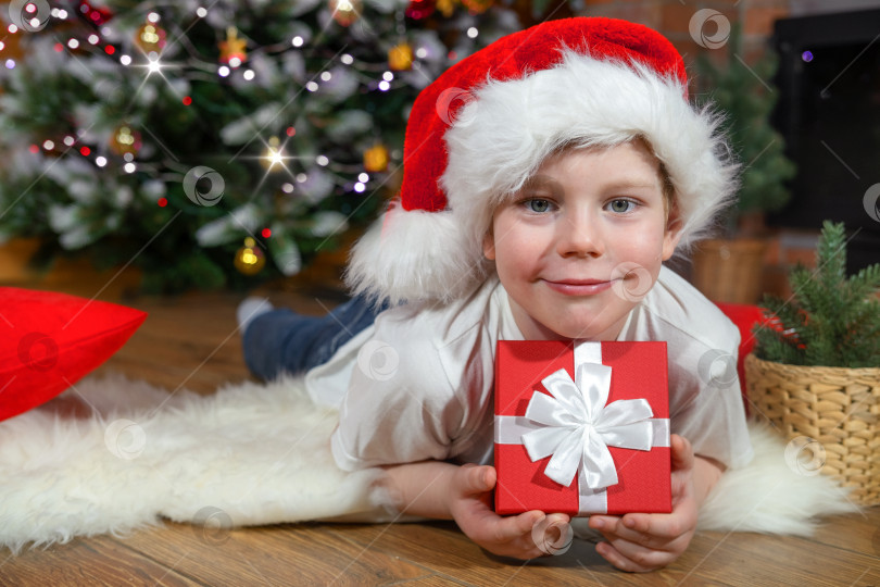 Скачать Счастливого Рождества, дети. Мальчик в шапке Санта-Клауса с подарком от Санта-Клауса, возле рождественской елки с рождественскими гирляндами и камином. Семейное Рождество. Баннер фотосток Ozero