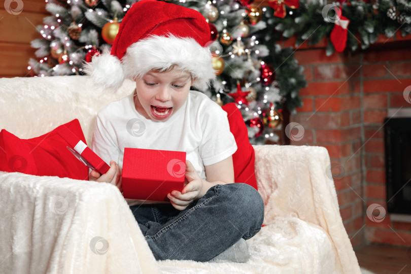 Скачать Счастливого Рождества, дети. Эмоции ребенка. Радостный удивленный малыш-мальчик в шляпе Санты открыл рождественскую коробку с подарком от Санта-Клауса возле рождественской елки с гирляндами, возле камина. Семейное Рождество. фотосток Ozero