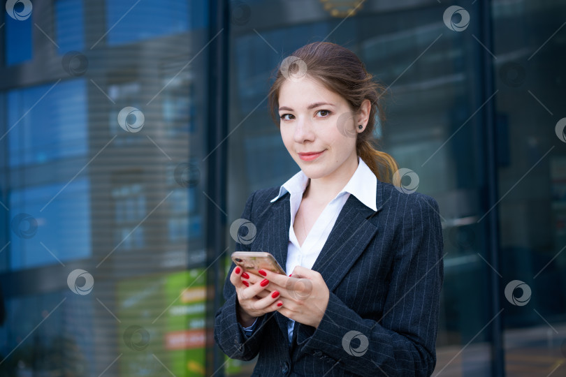 Скачать красивая молодая девушка-брюнетка в рубашке и пиджаке стоит на фоне офисного здания с телефоном в руках в солнечный день фотосток Ozero