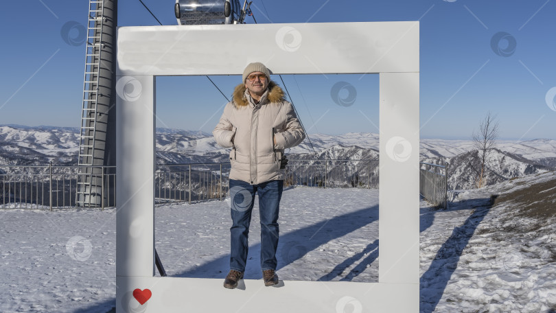 Скачать Мужчина в пуховике и шляпе стоит в рамке для фотографирования и улыбается. фотосток Ozero