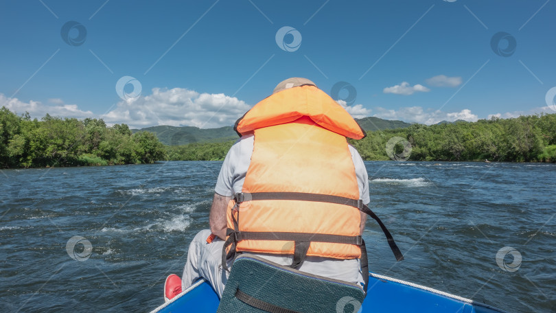 Скачать Мужчина в оранжевом спасательном жилете сидит на корме лодки, плывущей по реке фотосток Ozero