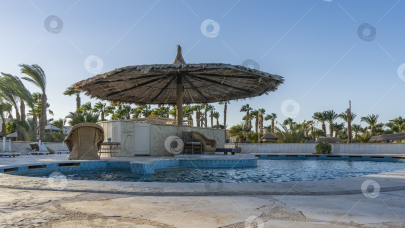 Скачать У бассейна с голубой водой стоят большой зонт от солнца и плетеные кресла. фотосток Ozero
