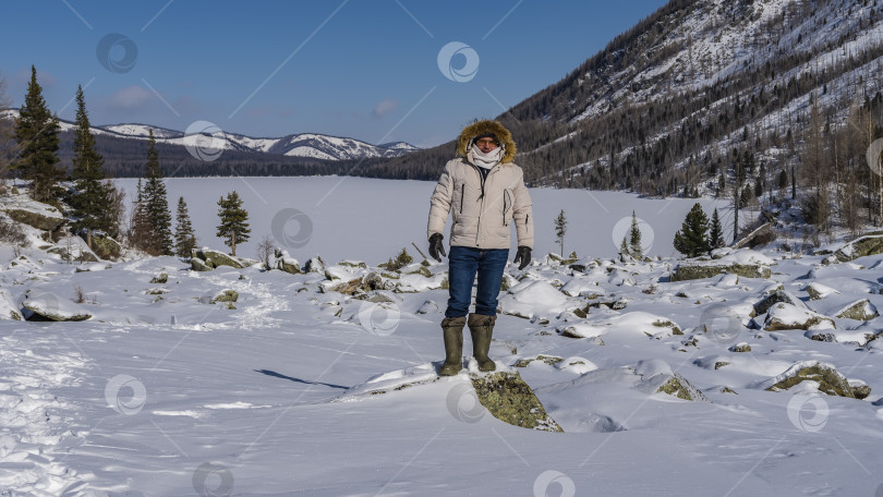 Скачать Мужчина в пуховике, меховом капюшоне, шарфе стоит в заснеженной долине на валуне.= фотосток Ozero