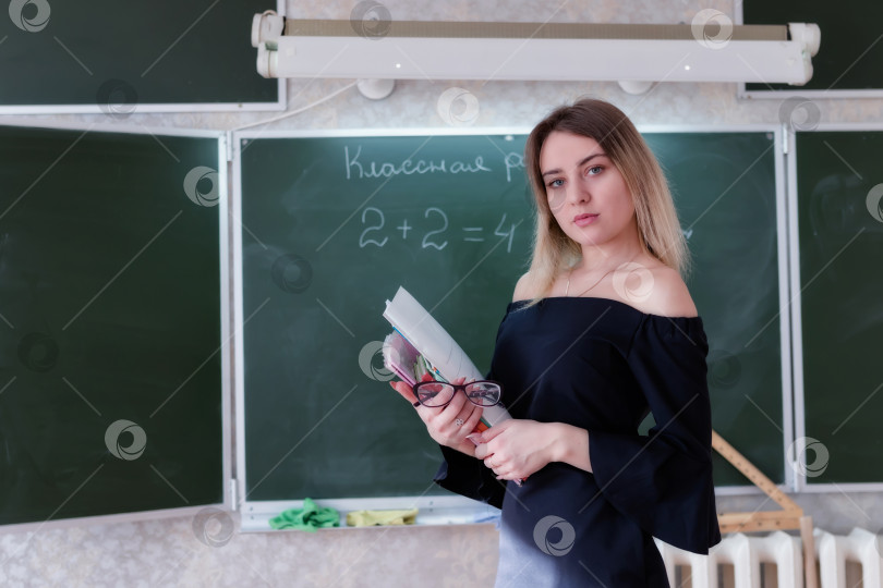 Скачать молодая очаровательная блондинка-учительница стоит в классе со стопкой учебников и тетрадей фотосток Ozero