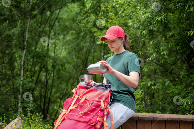 Скачать Туристка отдыхает после пешего похода по горам. Молодая туристка с красным рюкзаком пьет чай из термоса в горах. Пространство для копирования фотосток Ozero