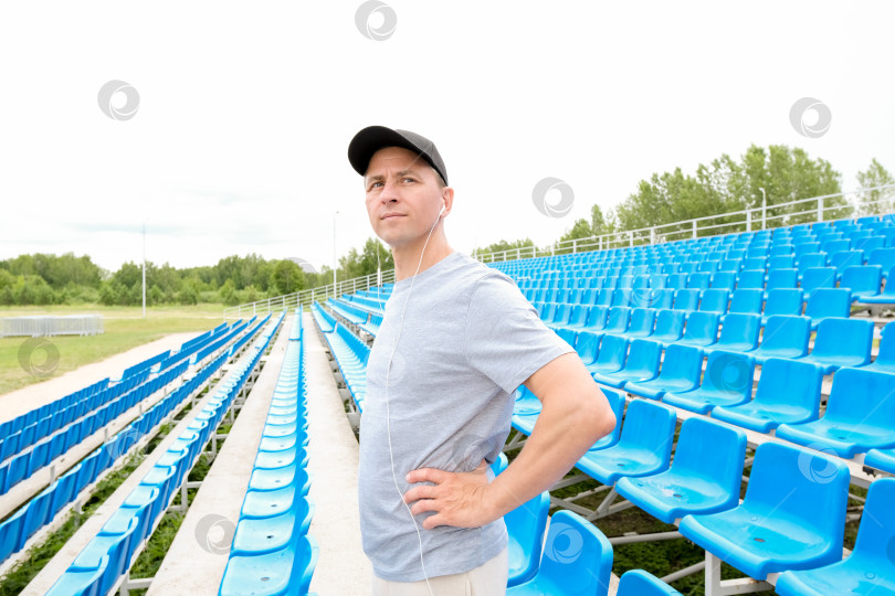 Скачать Мужчина в наушниках и черной кепке стоит на заднем плане стадиона и смотрит вперед. Концепция утренних физических упражнений на открытом воздухе фотосток Ozero