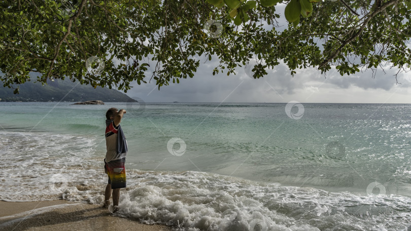 Скачать Мужчина стоит на пляже в полосе прибоя и смотрит вдаль на бирюзовый океан. фотосток Ozero