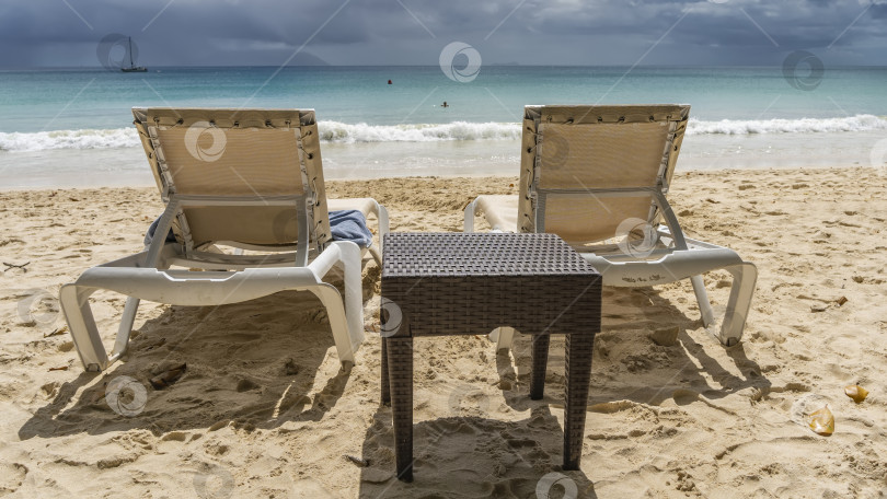 Скачать На песчаном пляже есть два шезлонга и плетеный столик. фотосток Ozero