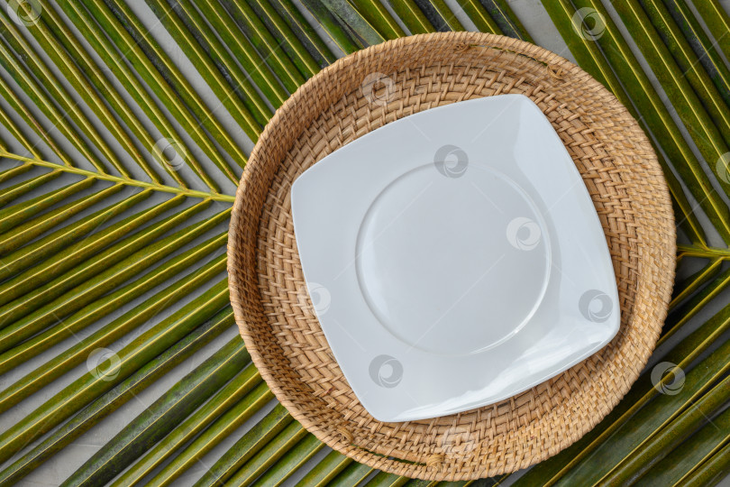 Скачать Пустая квадратная белая тарелка на аутентичном плетеном подносе на зеленом пальмовом листе. фотосток Ozero