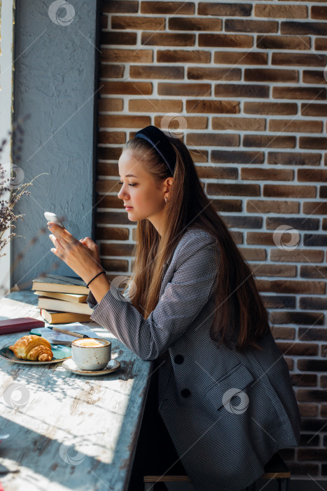 Скачать Молодая симпатичная девушка печатает на мобильном телефоне. Девушка пьет кофе в кофейне. фотосток Ozero