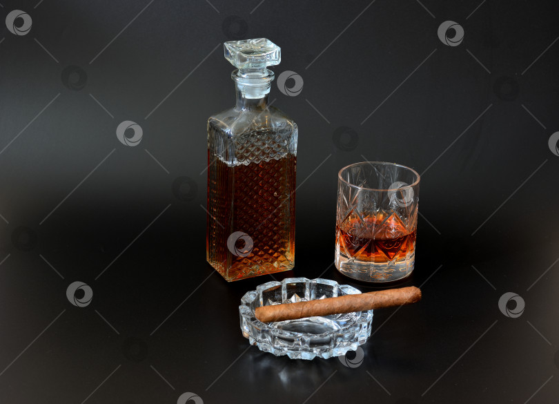 Скачать Хрустальный графин и граненый бокал для коньяка, а также стеклянная пепельница с кубинской сигарой на черном фоне. фотосток Ozero