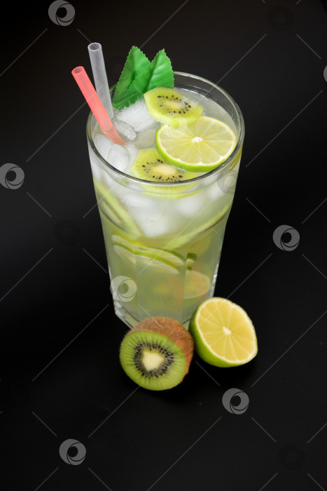 Скачать Высокий граненый стакан освежающего лимонада со льдом и соломинками на черном фоне, рядом с ним - спелая ветка и фрукт киви. фотосток Ozero