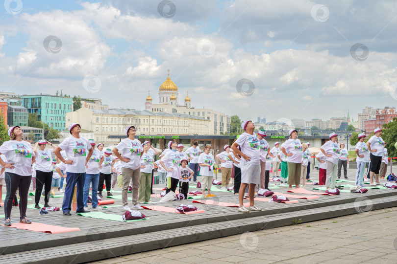 Скачать Московские пенсионеры в фирменных московских футболках и шапочках, участники программы «Московское долголетие, делают гимнастику в парке Музеон на фоне храма Христа Спасителя, голубого неба и красивых кучевых облаков. фотосток Ozero