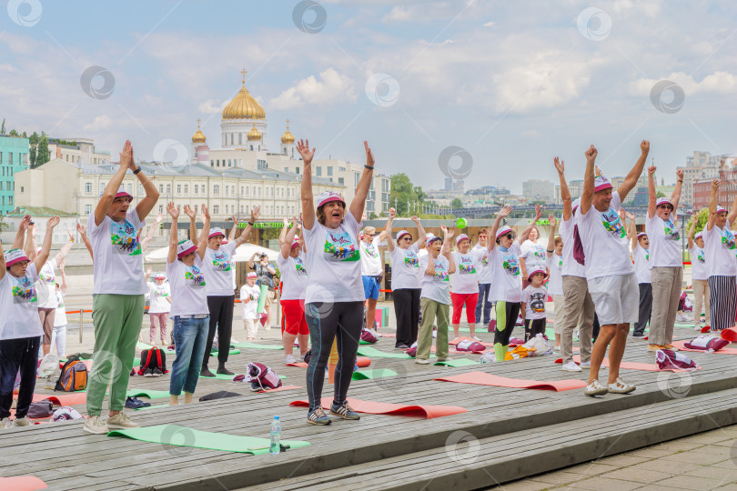 Скачать Московские пенсионеры в фирменных московских футболках и головных уборах делают гимнастику в парке Музеон на фоне храма Христа Спасителя, голубого неба и красивых облаков. фотосток Ozero